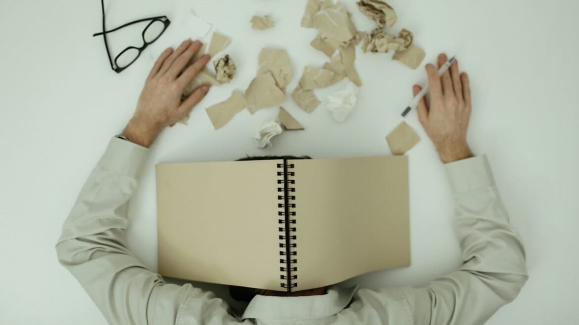Overweldigde man met stress en frustratie Man met hoofd op tafel en notitieboek over gezicht, omringd door verkreukeld papier, mentale overbelasting bij mannen